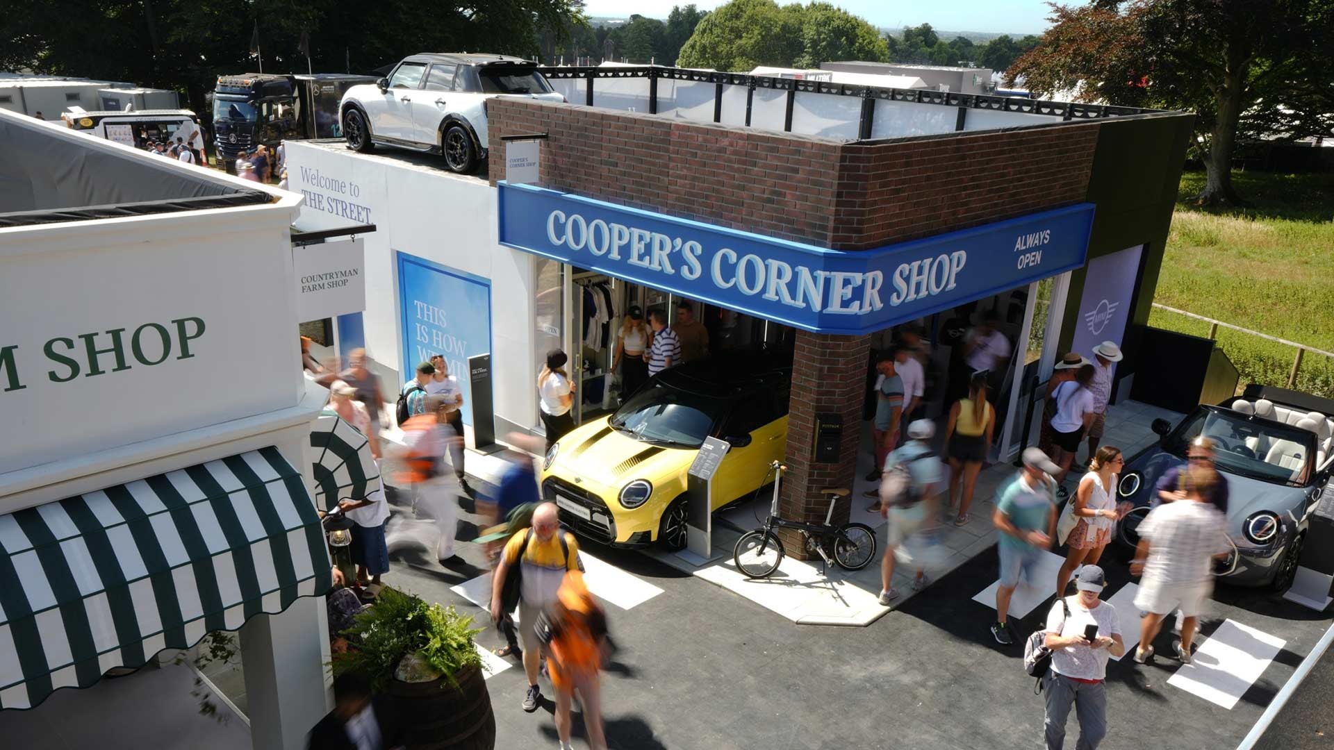 Crowd in motion at an outdoor car event, featuring a vibrant yellow car partially inside a “Cooper's Corner Shop” booth, with a bicycle and convertible nearby.